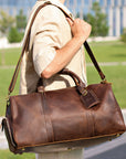 Brown vintage leather duffle bag with an adjustable strap carried over the shoulder, shown outdoors.