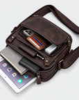 A dark brown leather crossbody bag shown open on a light gray background, filled with a tablet, phone, wallet, notebook, and other accessories.