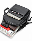 A black leather messenger bag shown open and partially filled with accessories, including a tablet, notebook, wallet, and books, on a clean white background.