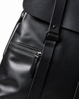 Close-up of a black full-grain leather backpack showing a front zip pocket, black strap, and buckle detail against a white background.