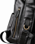 Close-up detail of a black leather backpack showing a vertical zippered pocket with a gold-tone zipper, a side pocket, and gold-tone metal D-ring hardware.