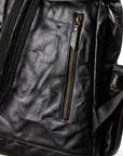 Close-up of a black leather backpack showing the front pocket with a vertical gold-tone zipper, textured leather surface, and part of the side strap on a white background.