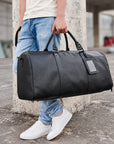 A person outdoors holding a large black leather duffel bag with dual top handles and a blank rectangular patch on the side. The person is wearing light blue jeans and white sneakers, standing beside a concrete pillar.