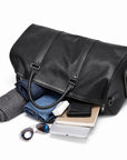 A black leather duffel bag shown open on a white background, with folded clothing, a wallet, a tablet, and sunglasses placed inside and around it.