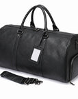 A black pebbled leather duffel bag with dual top handles, a zippered main compartment, a side pocket, a detachable shoulder strap, and a blank white luggage tag, shown on a white background.