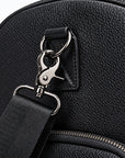 Close-up of a black pebbled leather bag with a black webbing strap attached via silver metal rings and a snap hook clasp, with a zipper visible below.