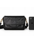 A black leather crossbody bag with gold-tone hardware, shown next to a black smartphone for scale.