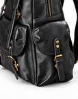 Close-up of a black leather backpack with gold-tone hardware, showing front flap pockets, buckle straps, and a zipper.