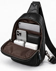 Black leather crossbody bag with an open front pocket showing a brown interior, filled with a smartphone, wallet, earbud case, and tissue. The bag has a black shoulder strap and silver-tone zippers, photographed on a clean white background.