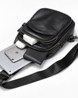 Black leather crossbody phone bag with a zippered front pocket, shown with a smartphone, earbuds, and a strap on a white background.