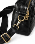 Close-up of a black leather crossbody bag with a wide black webbing strap and gold-tone hardware, including zippers and a ring clasp.