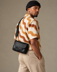 Rear view of a man wearing a black textured crossbody bag at his lower back, with an adjustable strap and brass-tone clips.