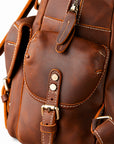 Close-up of a brown leather backpack showing the front pocket, stitching, buckle strap, and metal hardware.
