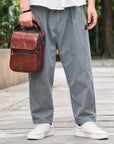 A person outdoors wearing loose gray pants and white sneakers, holding a brown leather crossbody bag. The background shows a blurred walkway and greenery.