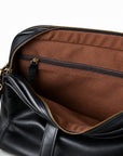 A close-up inside the black leather briefcase shows a horizontal zip pocket with a leather pull against brown lining.