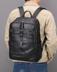 A man viewed from behind wearing a black leather backpack over a beige jacket and blue jeans, set against a solid gray studio background.