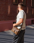 Man holding a brown leather bag against a brick wall.