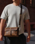 Man wearing sunglasses and a light gray t-shirt with a brown leather bag against a brick wall.