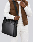 A man wearing a brown puffer vest, white long-sleeve shirt, and white pants carries a black leather messenger bag with a wide adjustable shoulder strap against a light gray studio background.