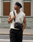 Man wearing a black leather crossbody bag with top handle, adjustable shoulder strap and gold-tone hardware.