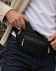 A black leather waist bag with multiple zippered compartments, shown being worn by a person in a beige jacket and blue jeans. The person is demonstrating the bag's capacity by inserting a black smartphone into its main compartment.