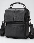 Black pebbled leather crossbody bag with a top handle, front zip pocket, and adjustable shoulder strap, photographed on a light gray studio background.