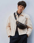 Man wearing a black leather sling bag across the chest with an adjustable strap and gold-tone zipper hardware.