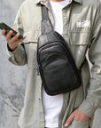 A man wearing a black leather crossbody phone bag across his chest, styled with an olive green shirt over a white t-shirt, holding a green smartphone against a gray concrete wall.