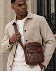 A man wearing a beige overshirt and white t-shirt carries a brown leather crossbody bag with a matching brown strap, standing in front of a stone building.
