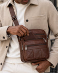 A man wearing a beige wool-blend jacket and white sweater carries a brown leather crossbody bag with multiple zippered compartments and a silver buckle.