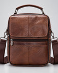 A brown textured leather crossbody bag with a top handle, front zip pocket, and matching shoulder strap, photographed against a light gray studio background.