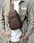 A man wearing a brown leather crossbody sling bag across his chest, styled with an olive green overshirt and white t-shirt against a textured gray wall.