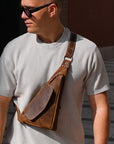 Man wearing a brown leather crossbody bag and sunglasses outdoors.