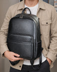 A man wearing a beige button-up shirt over a white t-shirt carries a black textured faux-leather backpack on his shoulder and holds it with one hand.