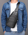 A black textured leather sling bag with a wide adjustable shoulder strap, worn crossbody over a blue denim shirt and white t-shirt, against a solid gray studio background.