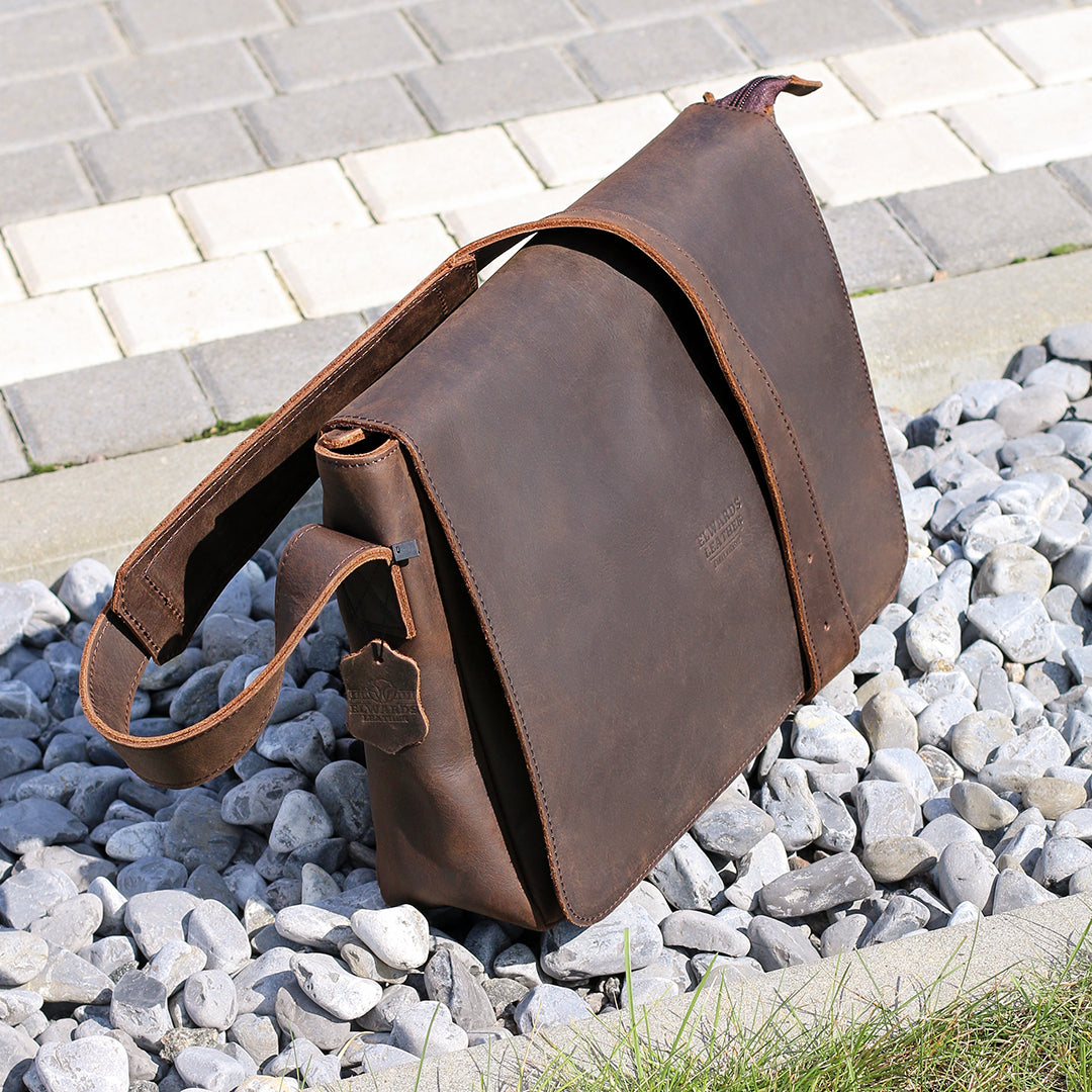 Angled view of a leather messenger bag placed on gravel, showing high-quality material and strap design