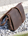 Angled view of a leather messenger bag placed on gravel, showing high-quality material and strap design