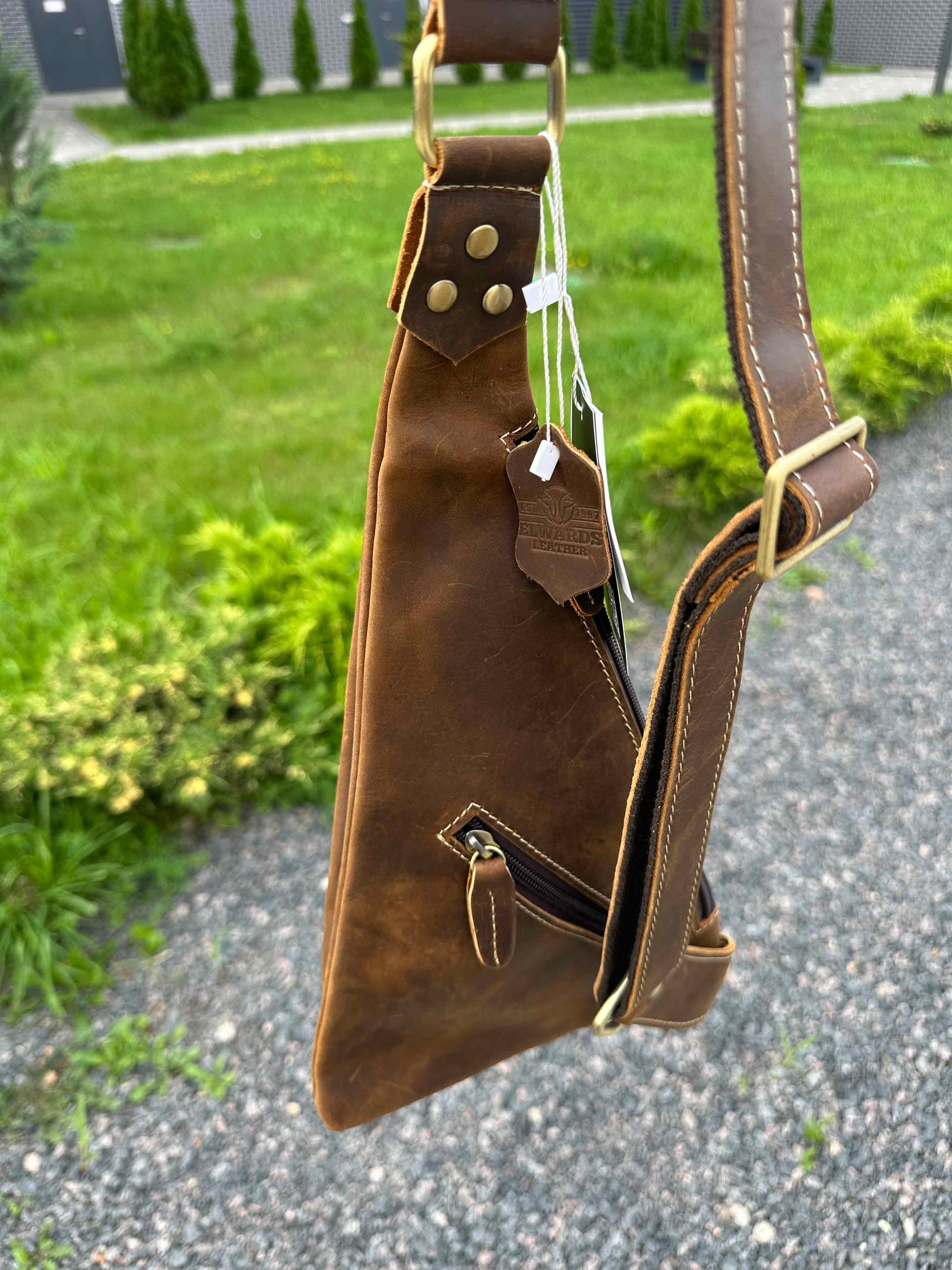 Brown leather crossbody bag with gold accents on a gravel path.