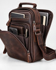 A dark brown distressed leather crossbody bag with multiple compartments, shown open to display its interior storage capacity with a smartphone, wallet, and tablet inside, set against a plain white studio background.