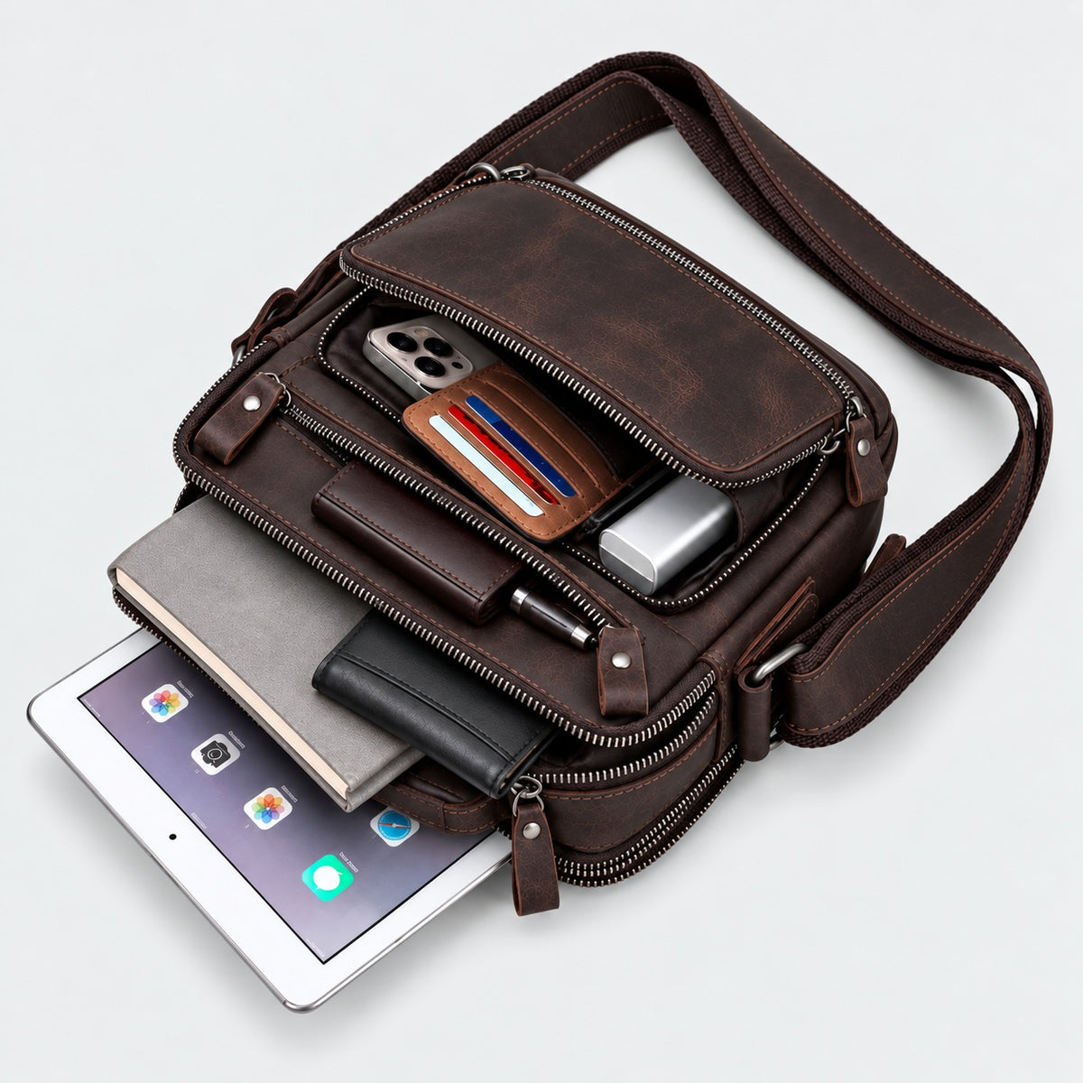 A dark brown leather crossbody bag shown open on a light gray background, filled with a tablet, phone, wallet, notebook, and other accessories.