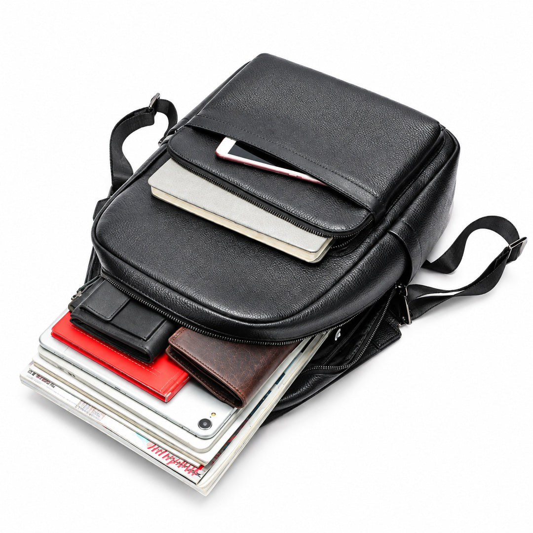 A black leather messenger bag shown open and partially filled with accessories, including a tablet, notebook, wallet, and books, on a clean white background.