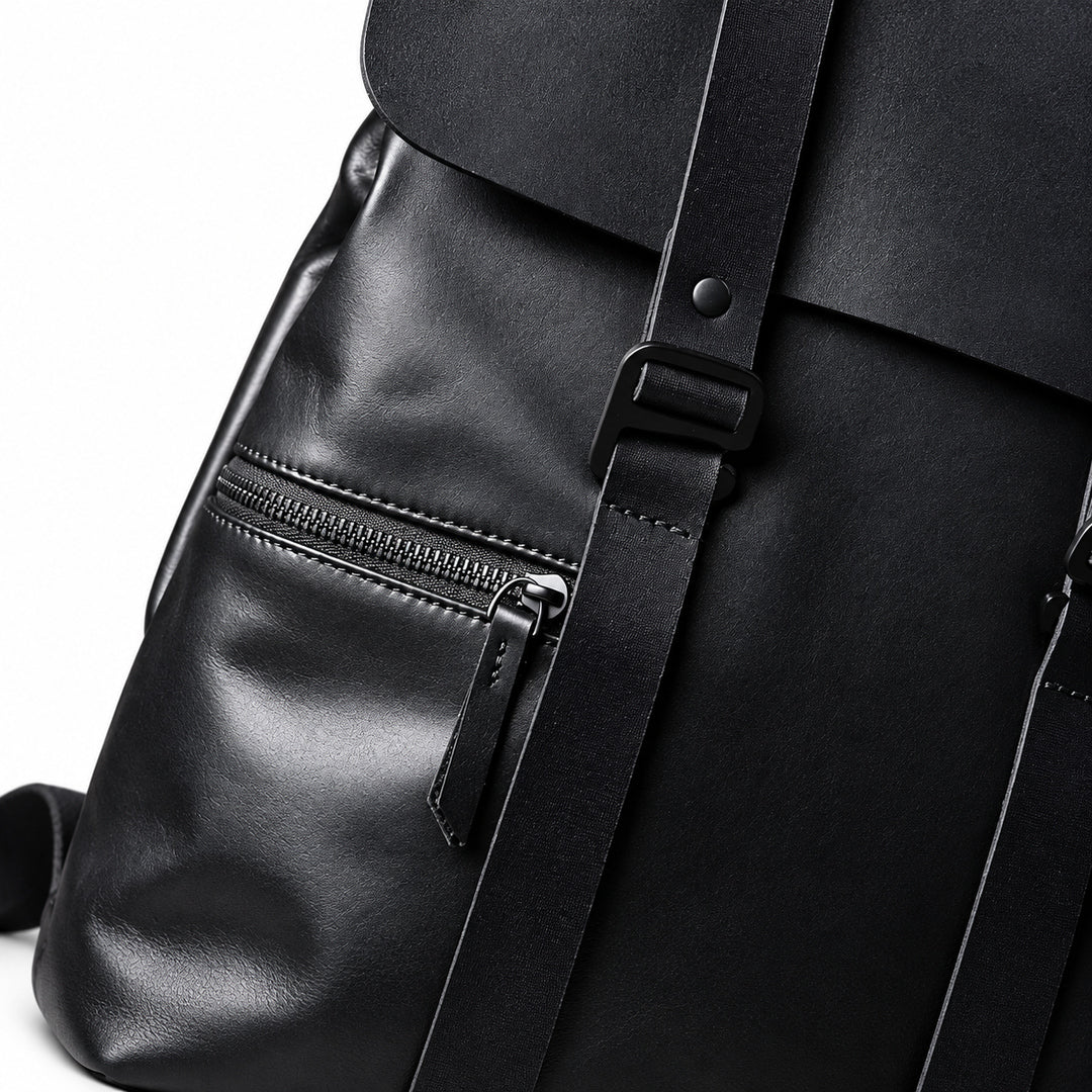 Close-up of a black full-grain leather backpack showing a front zip pocket, black strap, and buckle detail against a white background.