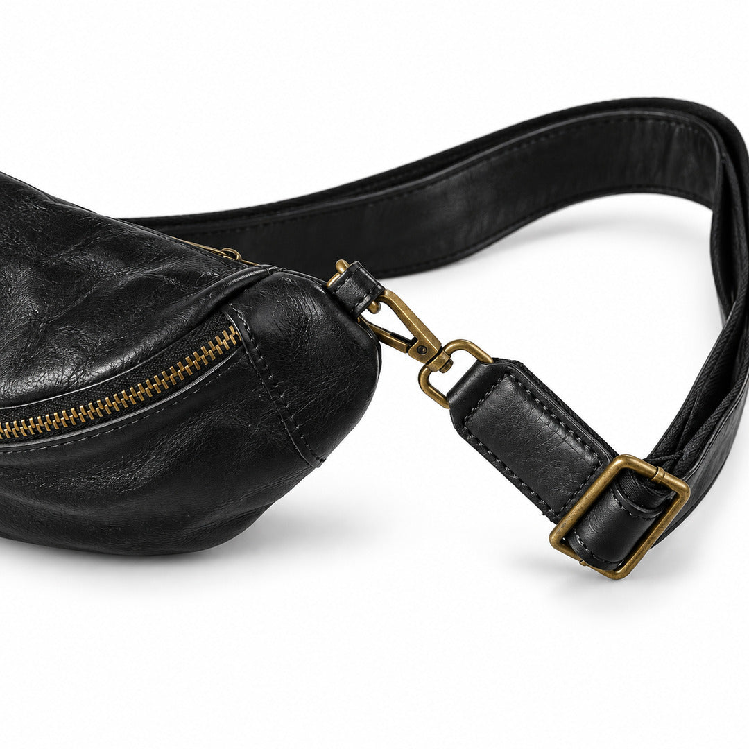 Close-up of a black leather sling bag with a gold-tone zipper and adjustable leather strap with brass buckle.