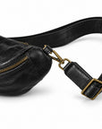 Close-up of a black leather sling bag with a gold-tone zipper and adjustable leather strap with brass buckle.