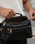 Close-up of a black leather crossbody bag opened to show the zippered main compartment, interior pocket and gold-tone hardware.