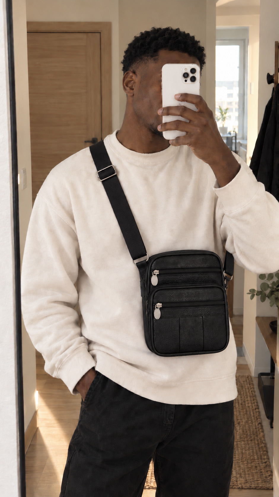 A man takes a mirror selfie wearing a black leather crossbody bag with silver hardware, styled with a cream sweatshirt and black jeans.