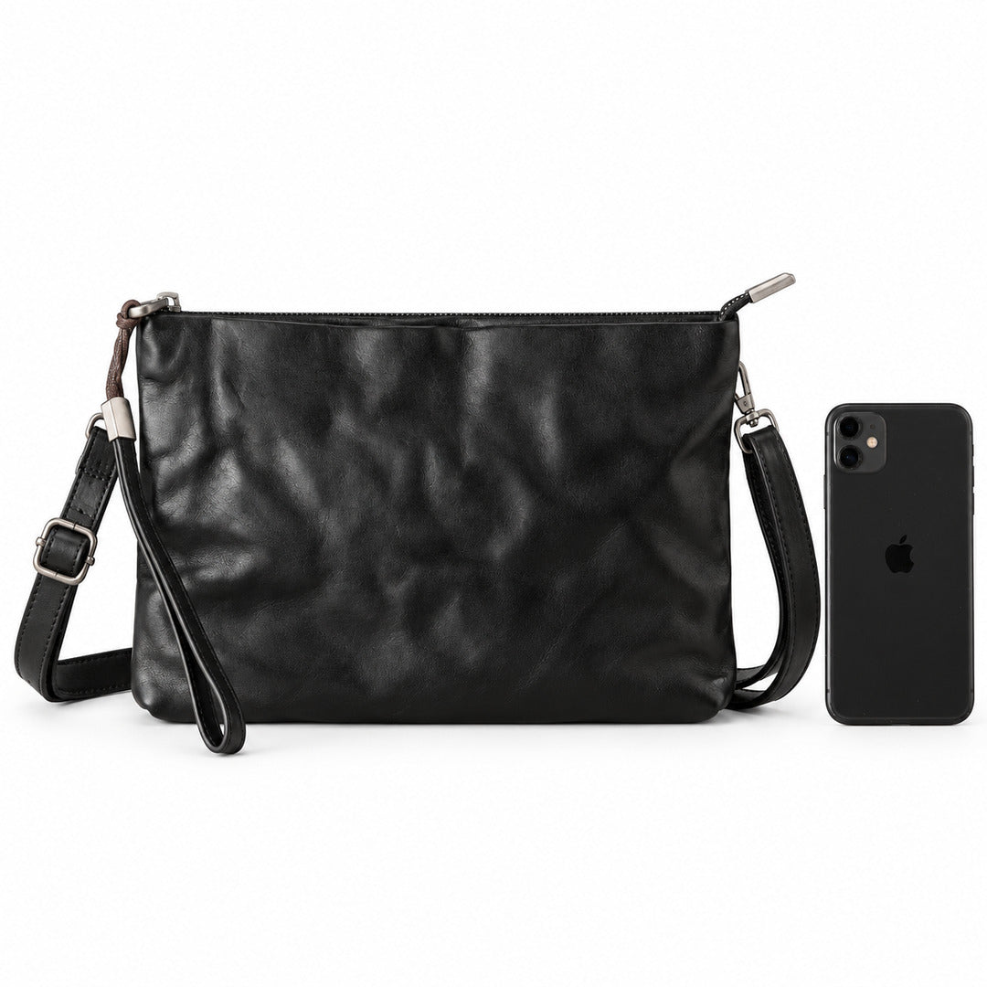 A black leather clutch bag with a wristlet strap and silver-tone hardware, shown next to a black smartphone for scale.