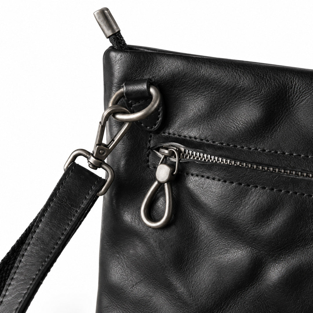 Close-up of a black leather crossbody bag showing a front zip pocket with a silver-tone keyring clasp and a black leather strap attached with metal hardware.
