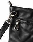Close-up of a black leather crossbody bag showing a front zip pocket with a silver-tone keyring clasp and a black leather strap attached with metal hardware.