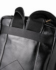 Close-up of a black faux-leather backpack showing the top handle, mesh ventilation panels, and a horizontal compression strap with gold-tone zipper pulls.